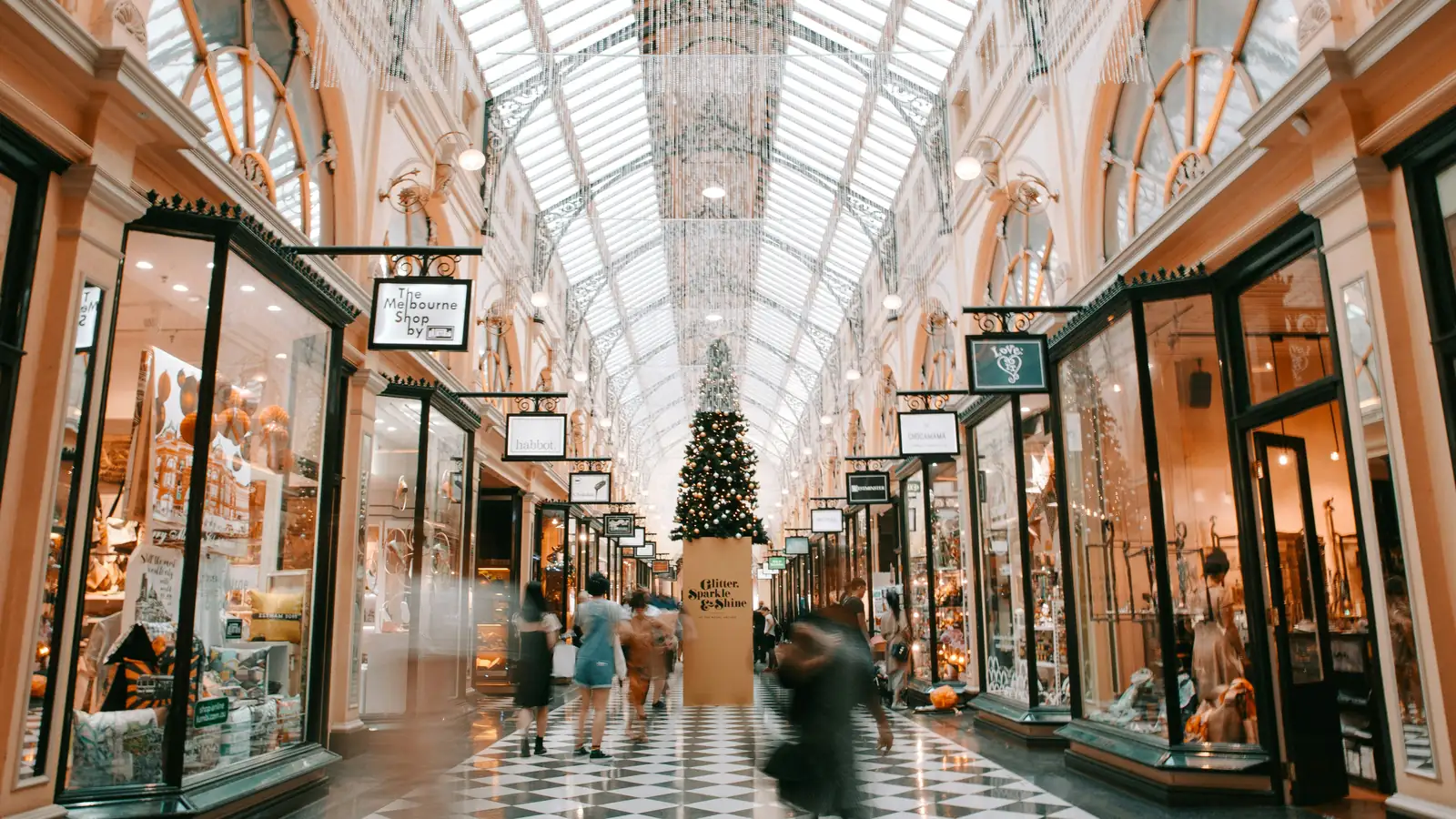A grand retail arcade under a glass roof, shoppers moving between lit storefronts